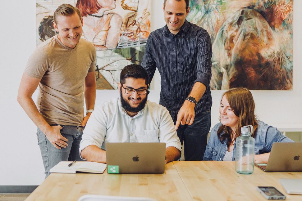 Four colleagues of diverse backgrounds smiling and gathered around a laptop at a light wood table, engaged in a collaborative working session in a bright office.
