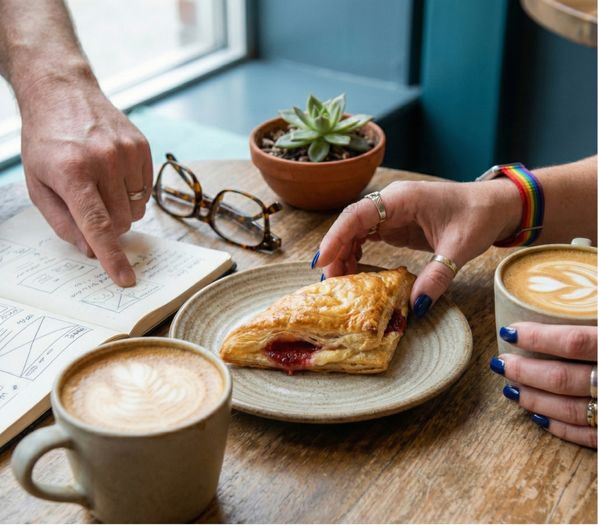 Close-up of two people at a wooden cafe table with lattes and a pastry; one person points to a hand-drawn website wireframe in a notebook while the other, wearing a rainbow watchband and blue nail polish, reaches for guava pastry.