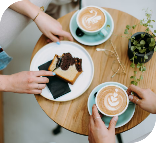 Top down view of two people seated at a small round table enjoying coffee and marble cake. There is a small plant on the table and some gold framed glasses. The coffees are decorated with beautiful late art.